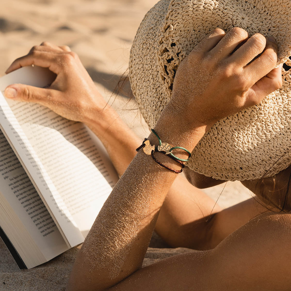 Woman reading a book on the beach, wearing a straw hat and bracelets, enjoying a summer day.